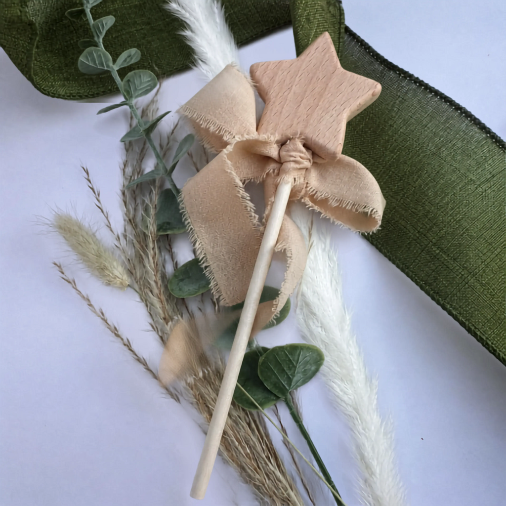A wooden star wand with a beige fabric bow and wooden stick, styled with dried grasses, green leaves, and a green ribbon on a light background.