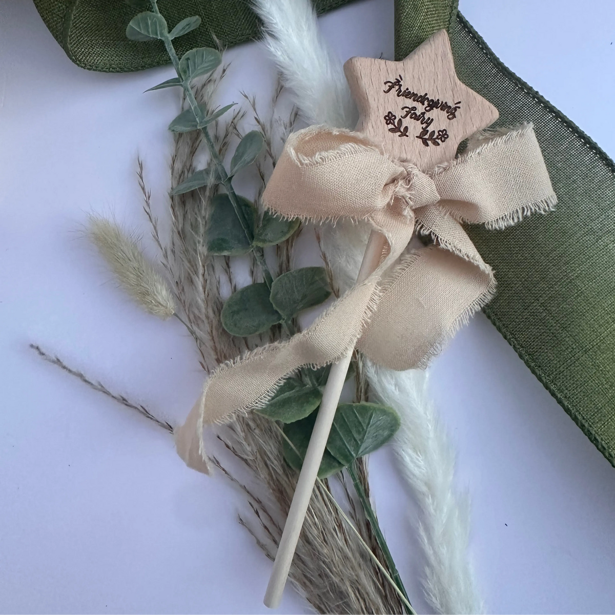 A wooden star-shaped keepsake wand with a beige frayed ribbon bow and a wooden stick, decorated with dried grasses, green eucalyptus leaves, and white fluffy stems on a light background.