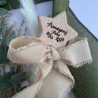 A close-up of a beige star-shaped wooden ornament with a matching frayed ribbon bow, featuring the engraved text 'Friendship Fairy' and small fairy illustrations. The ornament is styled with green burlap and dried grasses.