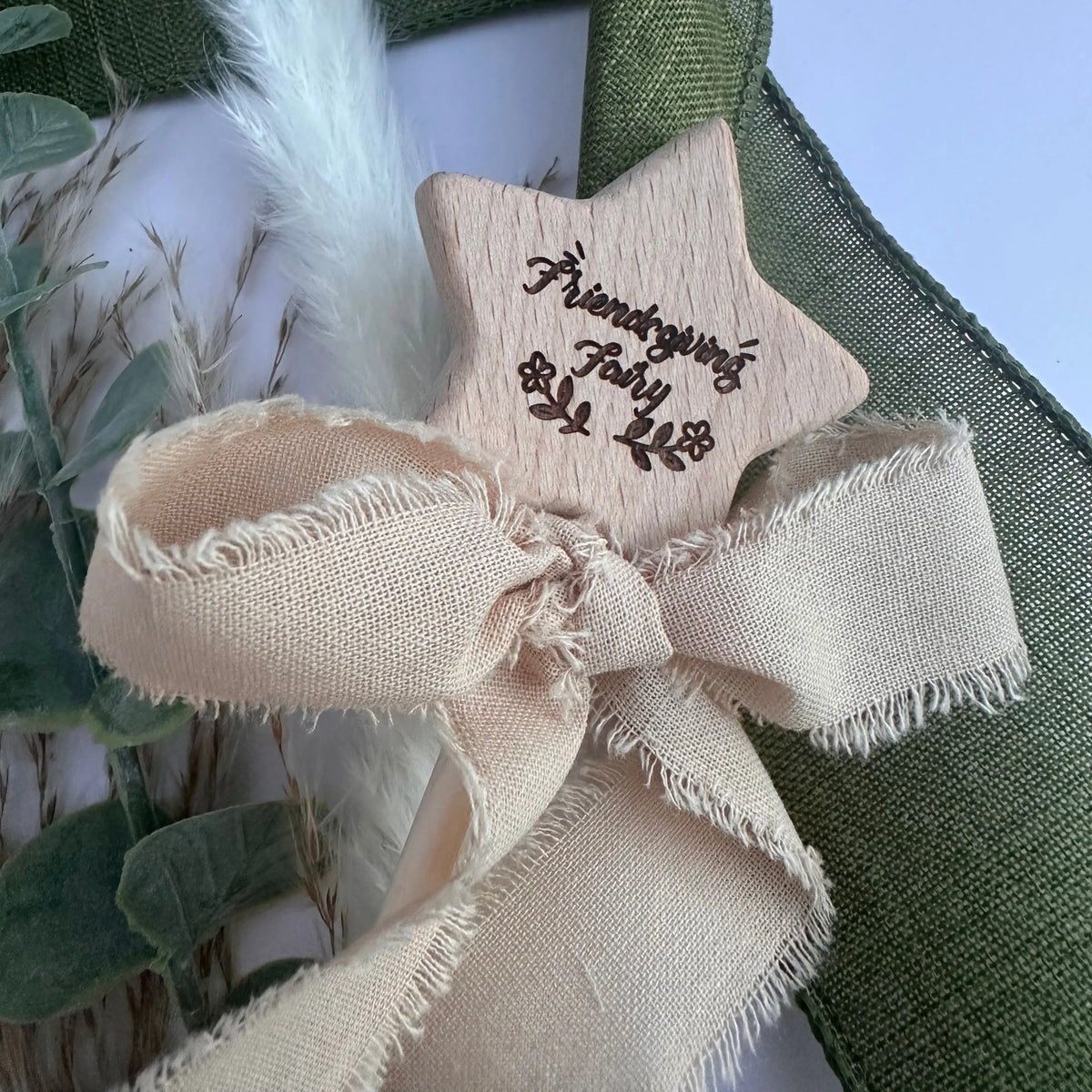 A close-up of a beige star-shaped wooden ornament with a matching frayed ribbon bow, featuring the engraved text 'Friendship Fairy' and small fairy illustrations. The ornament is styled with green burlap and dried grasses.