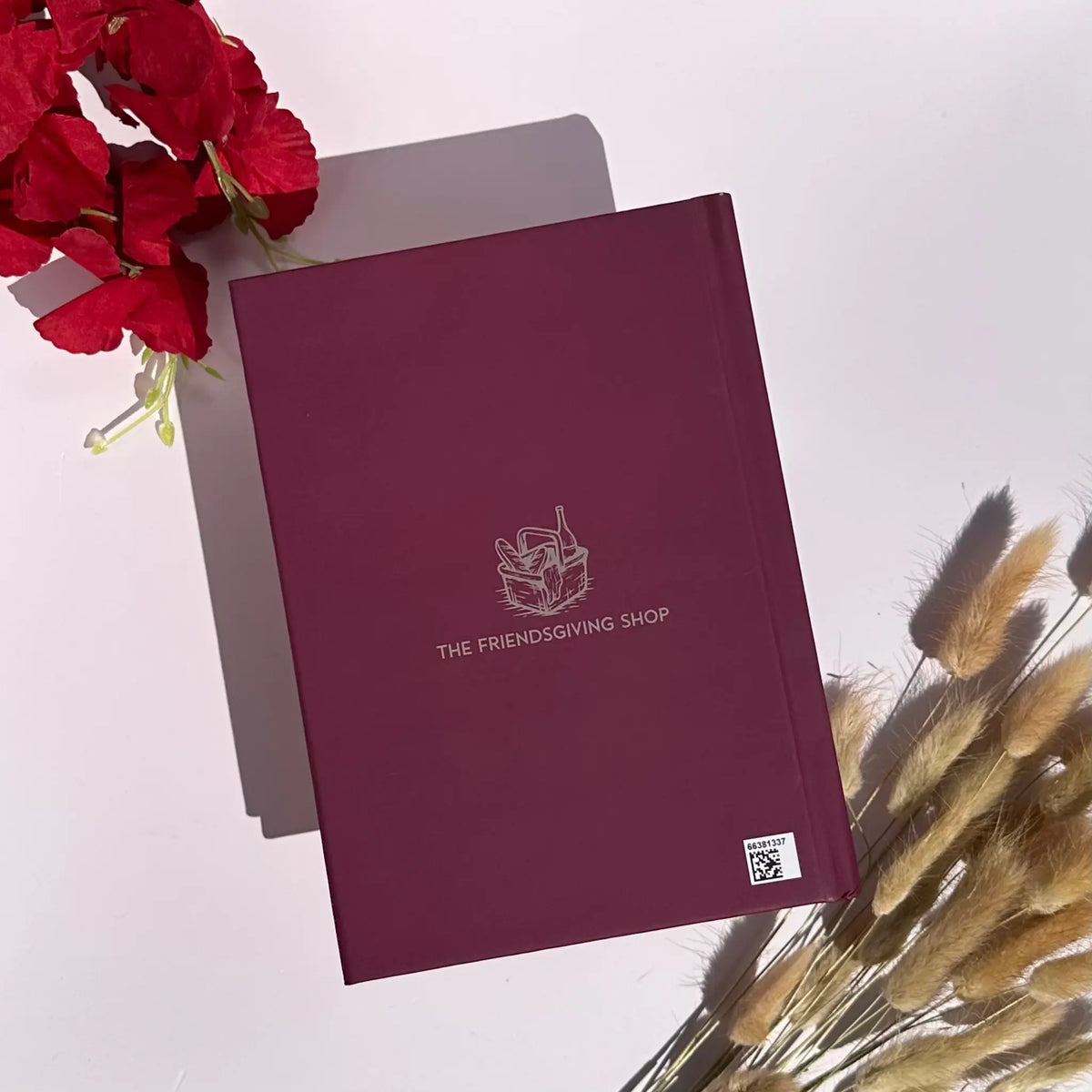 A hardcover notebook or journal in deep burgundy with a white line-art logo and the text 'THE FRIENDS GIVING SHOP' on the cover, styled on a white surface with red flowers and dried grass.