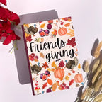 A hardcover notebook with a fall-themed cover featuring pumpkins, turkeys, leaves, and the words 'Friends giving' in black script, styled on a white surface with red flowers and dried grass.