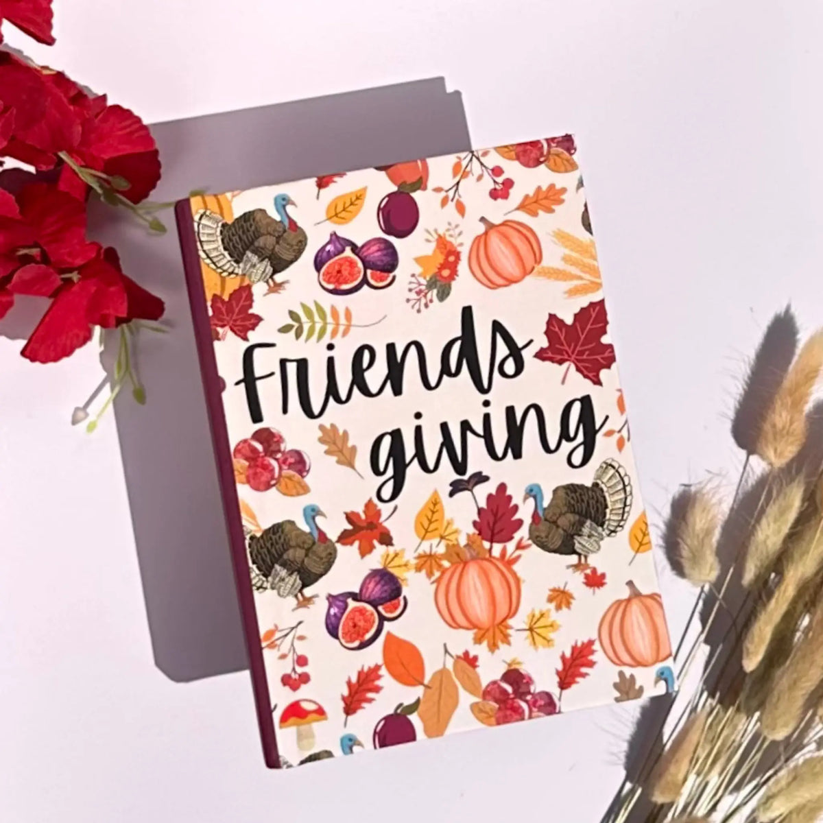 A hardcover notebook with a fall-themed cover featuring pumpkins, turkeys, leaves, and the words 'Friends giving' in black script, styled on a white surface with red flowers and dried grass.