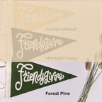 Three triangular pennant flags with the script word 'Friendsgiving' are shown in three colorways: Golden Wheat, Midnight Navy, and Forest Pine. The top two flags are shown faded and overlaid with their color names, while the bottom flag is in full color with the text 'Forest Pine' below it.