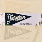 Three overlapping pennant flag designs labeled Friendlygivens, with color names Golden Wheat, Midnight Navy, and Forest Pine shown beneath them on a beige background with dried grass illustrations.