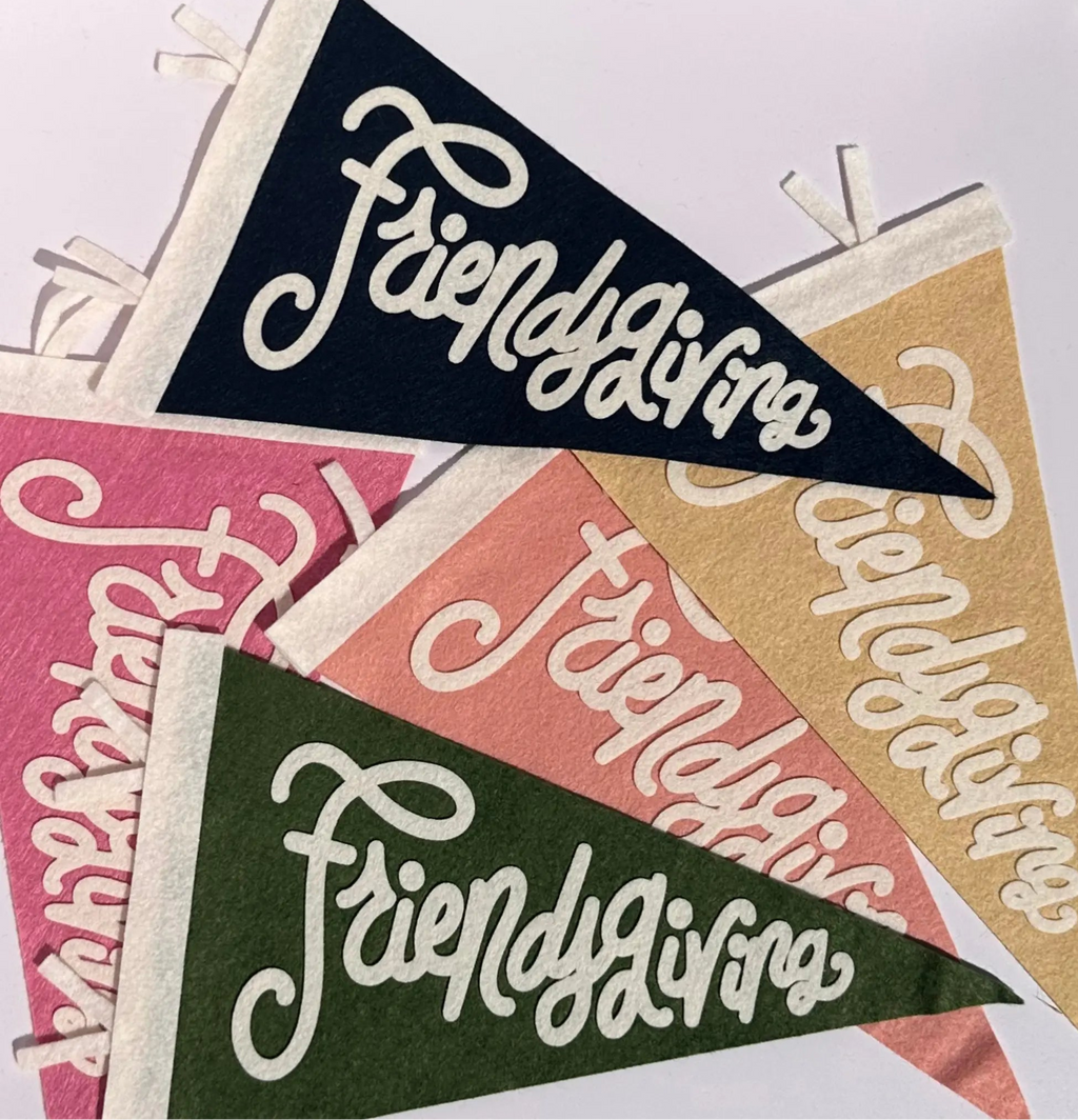 Four triangular felt pennants with white lettering reading 'Friendsgiving' in different colors: navy, pink, peach, and olive green, arranged on a white background.
