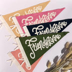 Four triangular felt pennants with white script reading 'Friendsgiving' are arranged diagonally on a light background, with dried grass stems in the lower right corner. The pennants are in tan, pink, navy, and olive green.