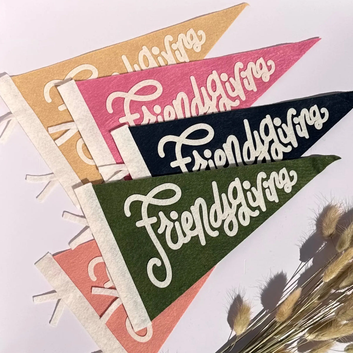 Four triangular felt pennants with white script reading 'Friendsgiving' are arranged diagonally on a light background, with dried grass stems in the lower right corner. The pennants are in tan, pink, navy, and olive green.