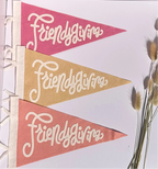 Three triangular pennant flags with the words 'Friendsgiving' in white script, shown in pink, tan, and coral colors, hanging vertically with white ribbons on the left side against a white background with dried grass stems on the right.