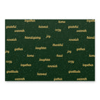 A square dark green fabric or paper product with a festive holiday pattern, featuring words like 'grateful', 'warmth', 'friendship', 'joy', 'harvest', and 'together' in gold script, accented with small white speckles. The item is shown flat against a clean white background with a subtle drop shadow.