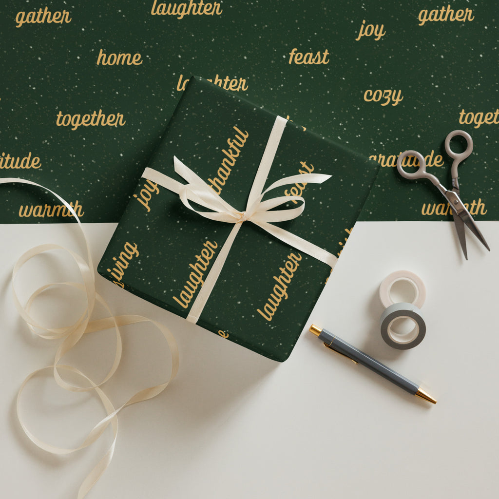 Dark green wrapping paper sheets with gold text and white speckles, shown wrapped around a gift box tied with a white ribbon, alongside scissors, washi tape, a pen, and loose ribbon on a white surface.