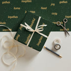 Dark green wrapping paper sheets with gold text and white speckles, shown wrapped around a gift box tied with a white ribbon, alongside scissors, washi tape, a pen, and loose ribbon on a white surface.