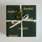A square gift box wrapped in dark green wrapping paper with gold script words and white speckles, tied with a white ribbon and decorated with a small green pine sprig, on a light gray background.