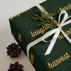 Dark green wrapping paper with gold script text and white snowflake speckles, shown on a gift box tied with a white ribbon and decorated with a small green sprig and pinecones.