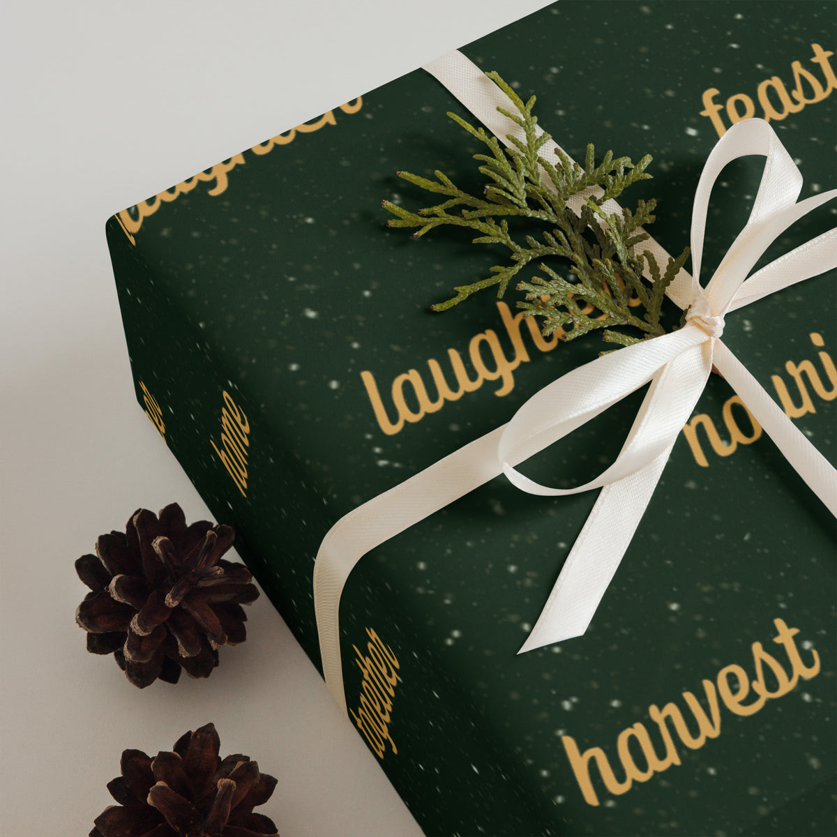 Dark green wrapping paper with gold script text and white snowflake speckles, shown on a gift box tied with a white ribbon and decorated with a small green sprig and pinecones.
