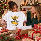 A woman holds up a white graphic t-shirt featuring a gold crown and the text 'King Mac n' Cheese!' above an illustration of a bowl of macaroni. The scene is set in a cozy, decorated Christmas home with another woman smiling in the background, surrounded by her wrapping station with gift boxes, wrapping paper, and holiday greenery.