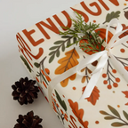A gift box wrapped in cream wrapping paper with a fall harvest pattern of orange leaves, brown berries, and green branches, tied with a white ribbon and topped with a small green sprig. Two pinecones rest on the white surface beside the box.