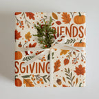 A square gift box wrapped in cream-colored paper with an autumn fall pattern featuring orange pumpkins, red and orange leaves, green branches, and the words 'FRIENDS' and 'GIVING'. The box is tied with a white satin ribbon and decorated with a small green pine sprig, set against a neutral light gray background.