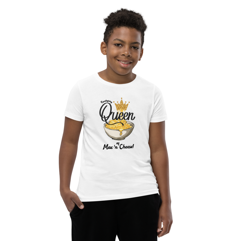 A young boy wearing a white graphic t-shirt with a 'Queen of Mac 'n Cheese' cereal design, paired with black pants, photographed against a solid white background.