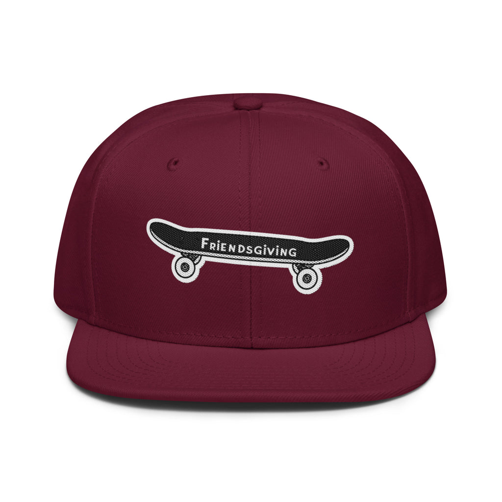 A maroon flat-brim snapback cap featuring an embroidered black skateboard with white wheels and the text 'Friendsgiving' on the front.