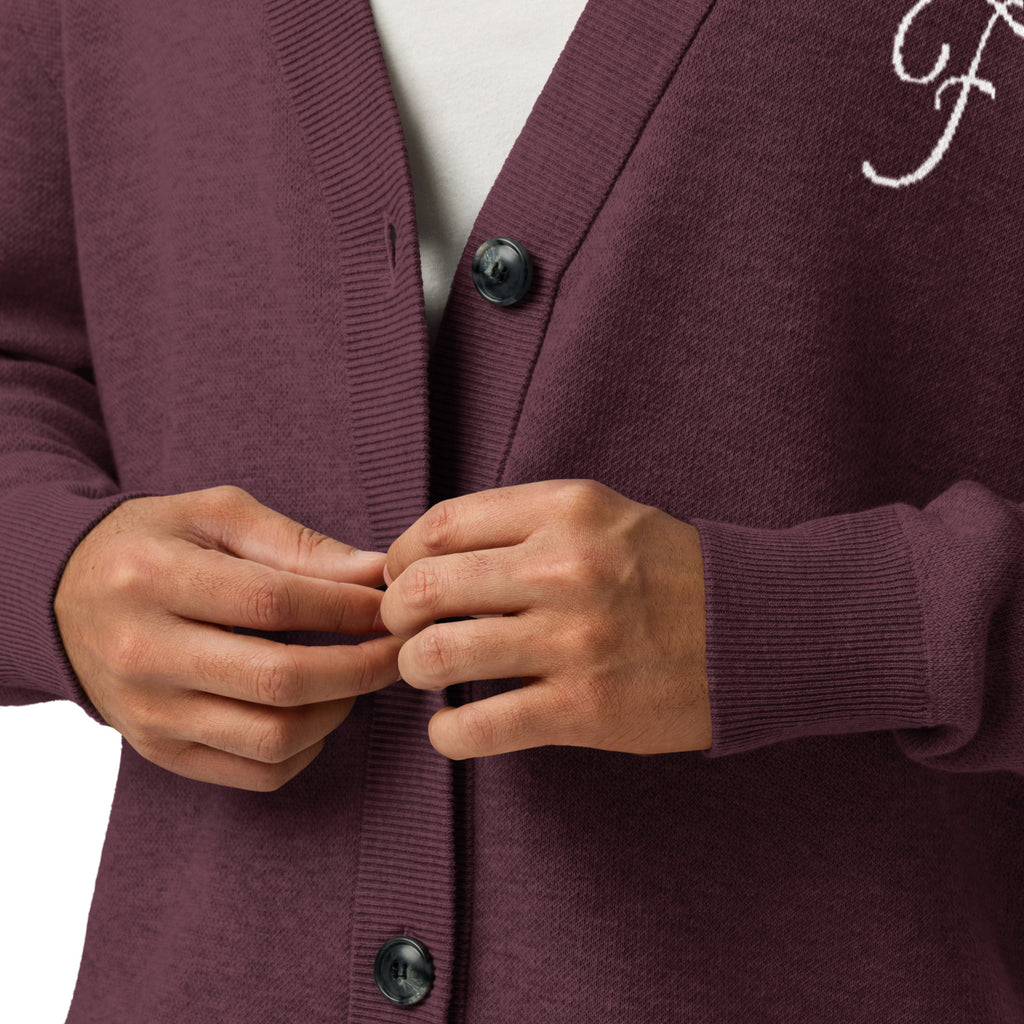 Close-up of a person wearing a ribbed cardigan in a muted plum-burgundy color, buttoning the front. A white undershirt is visible at the neckline, and a white embroidered script monogram appears on the upper left chest.