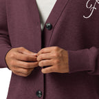 Close-up of a person wearing a ribbed cardigan in a muted plum-burgundy color, buttoning the front. A white undershirt is visible at the neckline, and a white embroidered script monogram appears on the upper left chest.