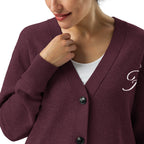 A close-up of a woman wearing a burgundy knit cardigan over a white top. The cardigan has a V-neck, black buttons, and a white embroidered script letter on the left chest.