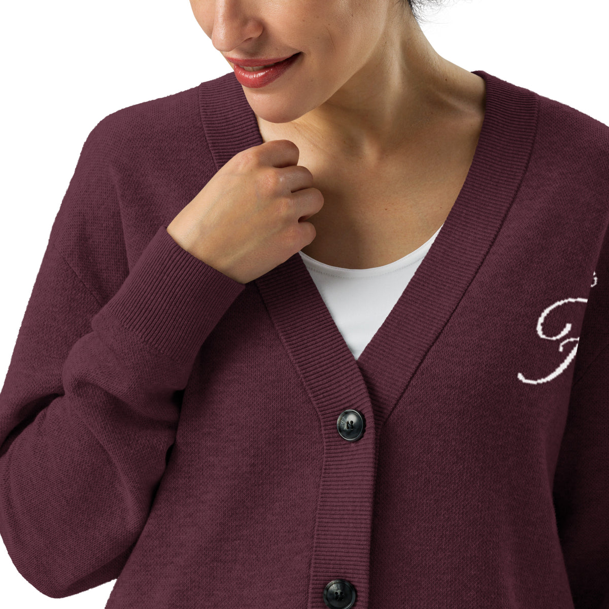 A close-up of a woman wearing a burgundy knit cardigan over a white top. The cardigan has a V-neck, black buttons, and a white embroidered script letter on the left chest.