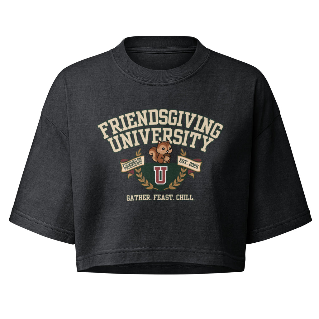 A dark charcoal gray cropped t-shirt featuring a vintage-style graphic on the front that reads 'FRIENDSGIVING UNIVERSITY' in cream lettering, with a brown teddy bear, a red and gold 'U' logo, and the phrase 'GATHER. FEAST.
