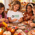 Four girls sitting around a table making beaded jewelry, with a girl in the center wearing a white graphic T-shirt that says 'FRIENDS GIVING' with a penguin logo. The table is decorated with autumn leaves, candles, and bowls of colorful beads.