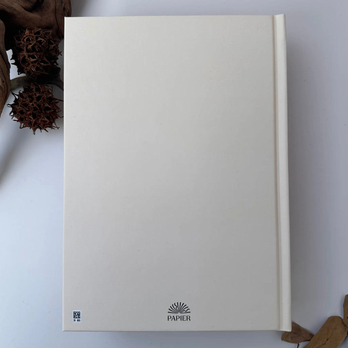 A cream-colored paper pad or notebook cover with the brand name 'PAPIER' printed at the bottom, photographed from above on a white surface with dried botanical props in the corners.