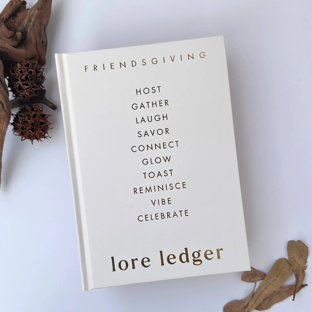 A hardcover book titled 'FRIENDSGIVING LORE LEDGE,' featuring a minimalist cream cover with black and gold typography, styled on a white background with dried botanical elements.