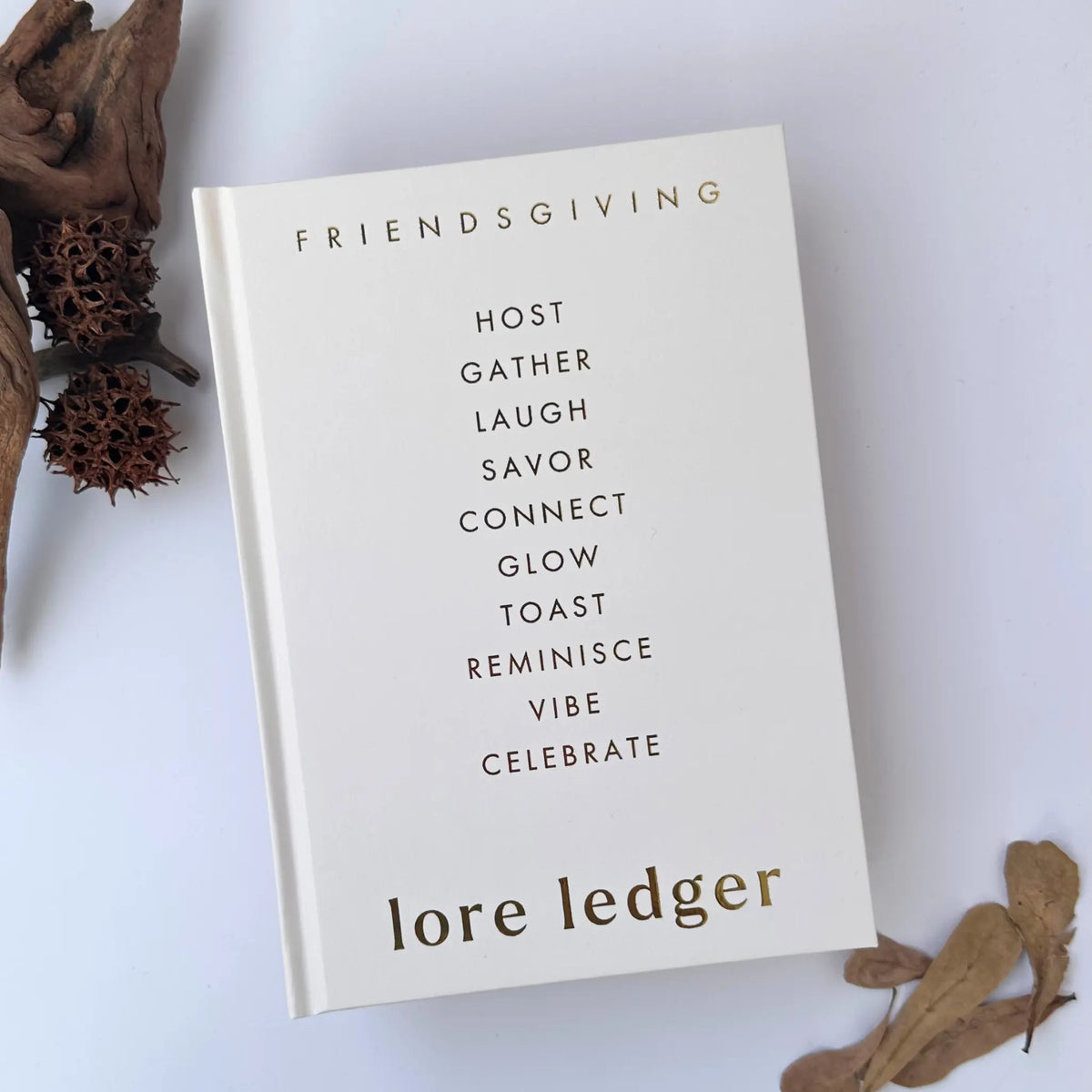 A hardcover book titled 'FRIENDSGIVING LORE LEDGE,' featuring a minimalist cream cover with black and gold typography, styled on a white background with dried botanical elements.