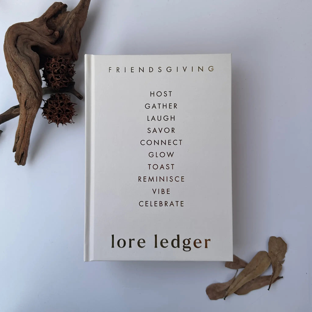 A hardcover book titled 'Friendsgiving lore ledger,' featuring a minimalist cream cover with black text, styled on a light gray background with dried botanicals and driftwood.