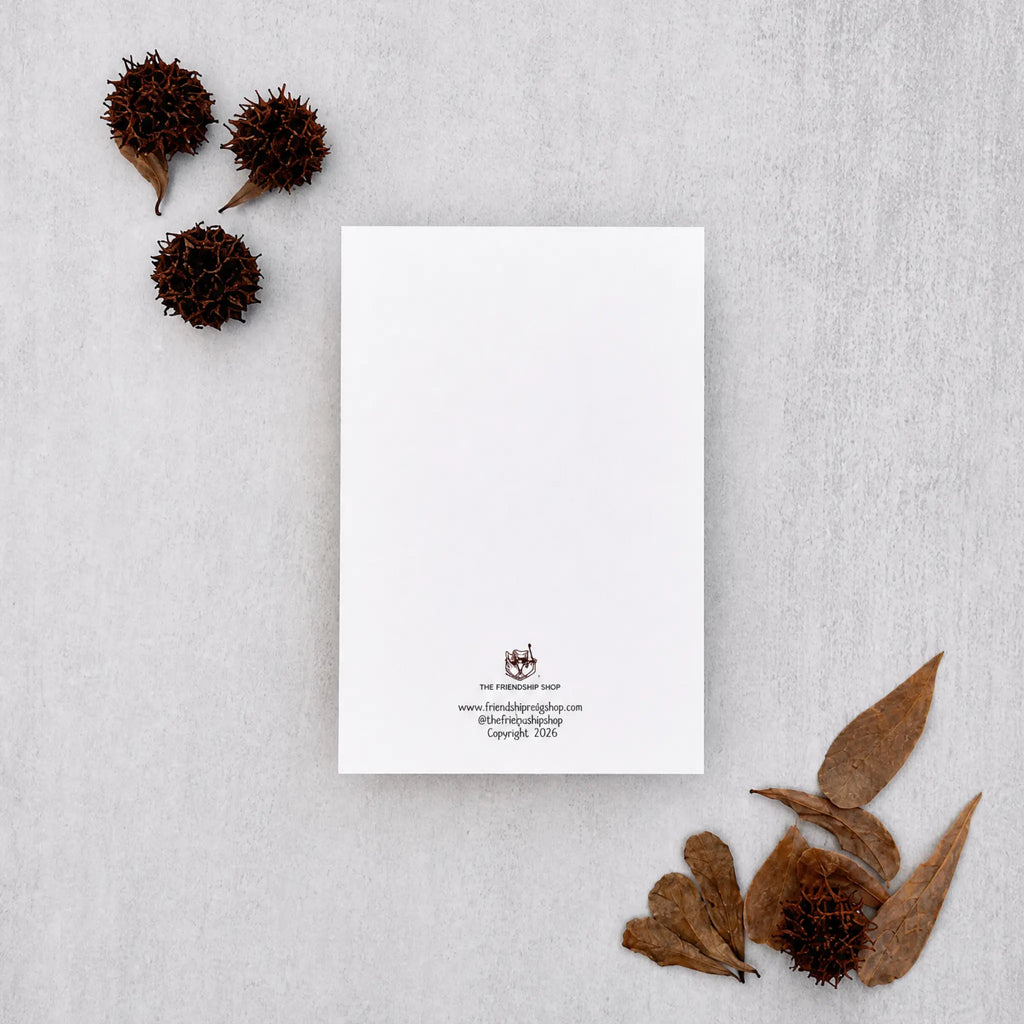 A minimalist white letterhead card with a small crest and contact details at the bottom, styled in a flat lay on a light gray textured surface with dried brown leaves and seed pods.