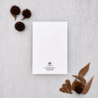A minimalist white letterhead card with a small crest and contact details at the bottom, styled in a flat lay on a light gray textured surface with dried brown leaves and seed pods.
