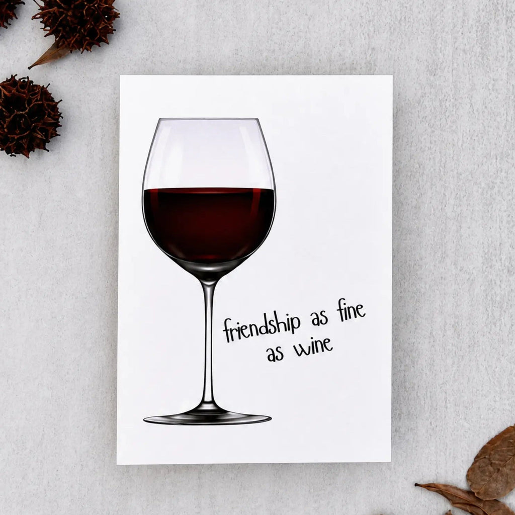 A white greeting card featuring an illustration of a wine glass with red wine and the handwritten-style text 'friendship as fine as wine.' The card is photographed on a light gray textured surface with dried brown leaves and seed pods arranged around it.