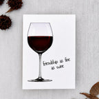 A white greeting card featuring an illustration of a wine glass with red wine and the handwritten-style text 'friendship as fine as wine.' The card is photographed on a light gray textured surface with dried brown leaves and seed pods arranged around it.
