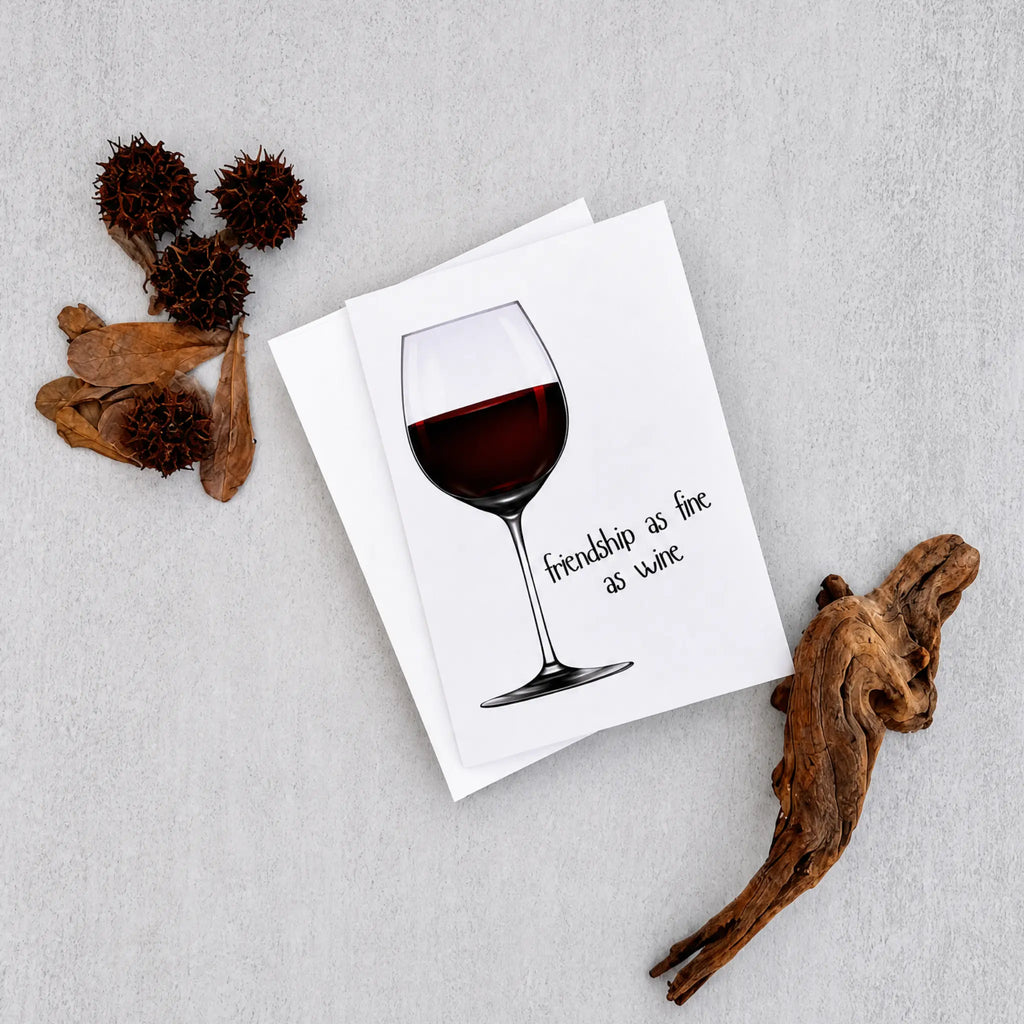 A white greeting card featuring an illustration of a wine glass filled with red wine and the handwritten phrase 'friendship as fine as wine', styled on a light gray textured background with dried botanical props.