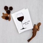 A white greeting card featuring an illustration of a wine glass filled with red wine and the handwritten phrase 'friendship as fine as wine', styled on a light gray textured background with dried botanical props.