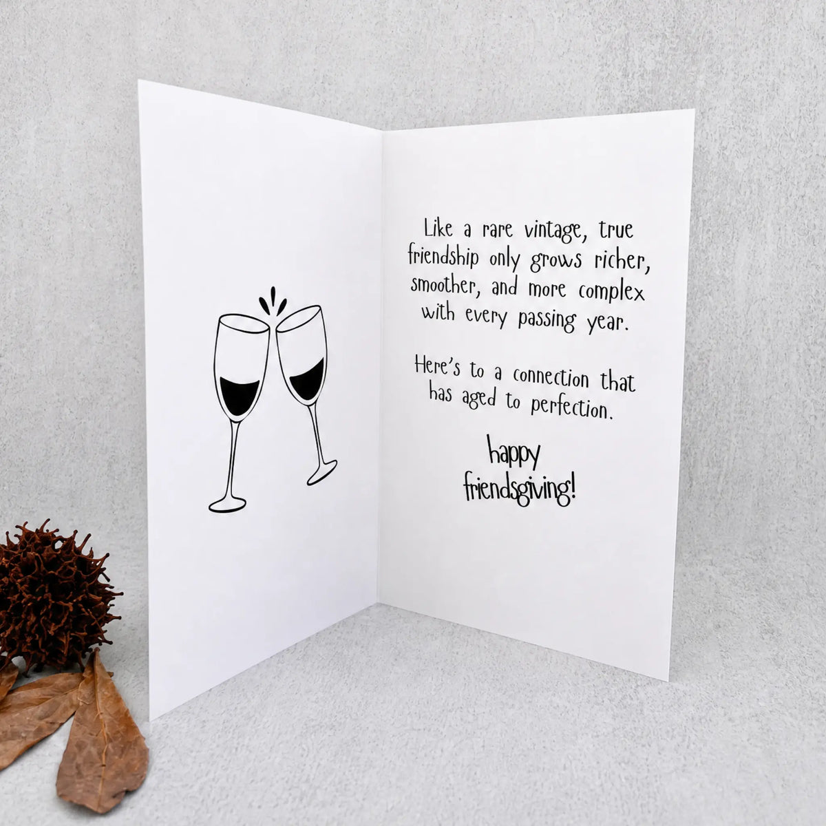 A white folding greeting card standing open on a light gray textured surface, with a black line drawing of two clinking wine glasses on the left panel and a handwritten-style message on the right panel. A brown pine cone and dried leaf sit to the lower left as props.