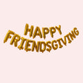Gold foil balloon letter banner spelling 'HAPPY FRIENDSGIVING' on a pale pink background.