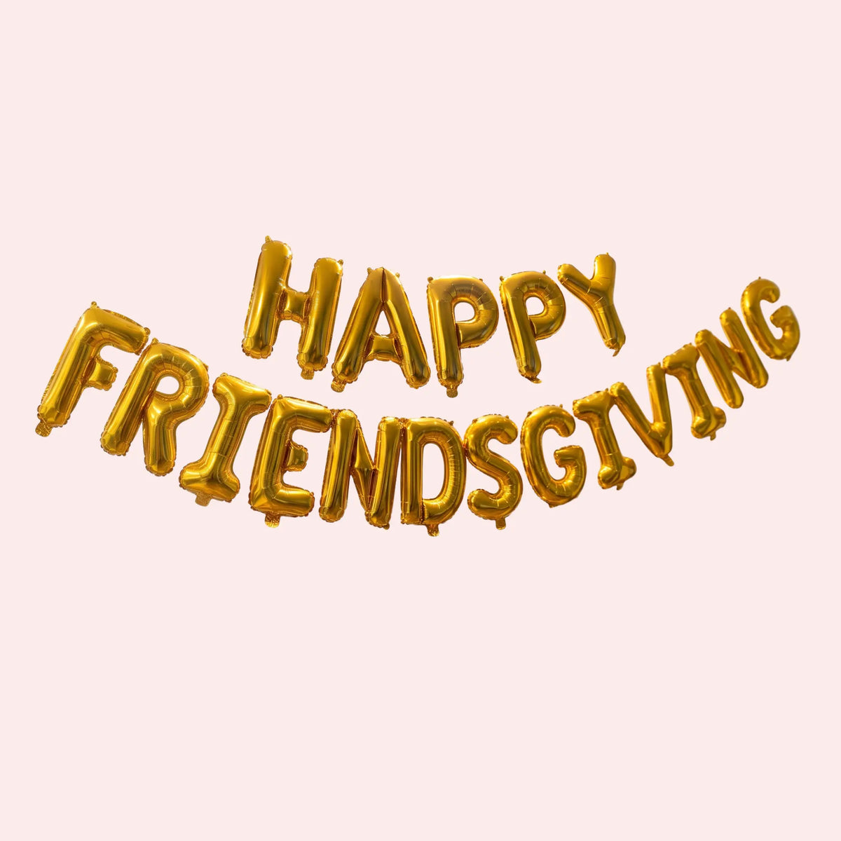 Gold foil balloon letter banner spelling 'HAPPY FRIENDSGIVING' on a pale pink background.
