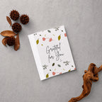 A square white greeting card with the phrase 'Grateful for You' in black script, decorated with small pink flowers, green leaves, and delicate botanical illustrations. The card is displayed in a flat-lay arrangement on a textured gray background, surrounded by natural decorative props including dried brown leaves, pinecones, and a piece of driftwood.