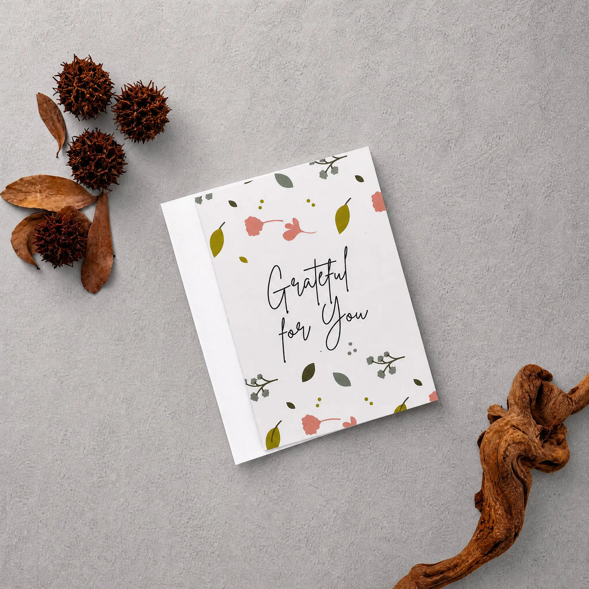 A square white greeting card with the phrase 'Grateful for You' in black script, decorated with small pink flowers, green leaves, and delicate botanical illustrations. The card is displayed in a flat-lay arrangement on a textured gray background, surrounded by natural decorative props including dried brown leaves, pinecones, and a piece of driftwood.