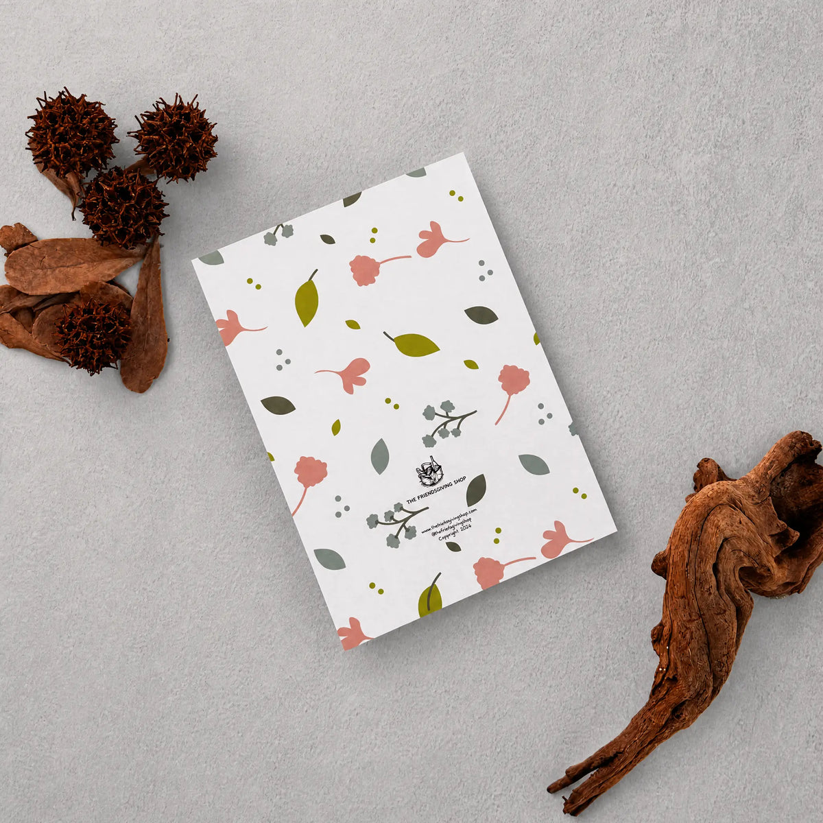 A flat rectangular printed card or notebook cover with a white background and a scattered floral pattern in muted pink, olive green, and gray, photographed on a light gray surface with dried botanical props around it.