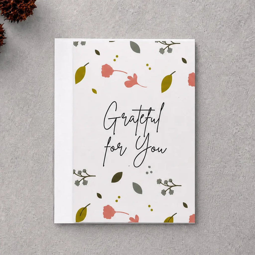 A square white greeting card with the phrase 'Grateful for You' in black script, decorated with small pink flowers, green leaves, and delicate botanical illustrations. The card is displayed in a flat-lay arrangement on a textured gray background, surrounded by natural decorative props including dried brown leaves, pinecones, and a piece of driftwood.