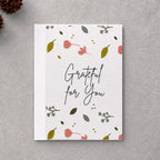 A square white greeting card with the phrase 'Grateful for You' in black script, decorated with small pink flowers, green leaves, and delicate botanical illustrations. The card is displayed in a flat-lay arrangement on a textured gray background, surrounded by natural decorative props including dried brown leaves, pinecones, and a piece of driftwood.