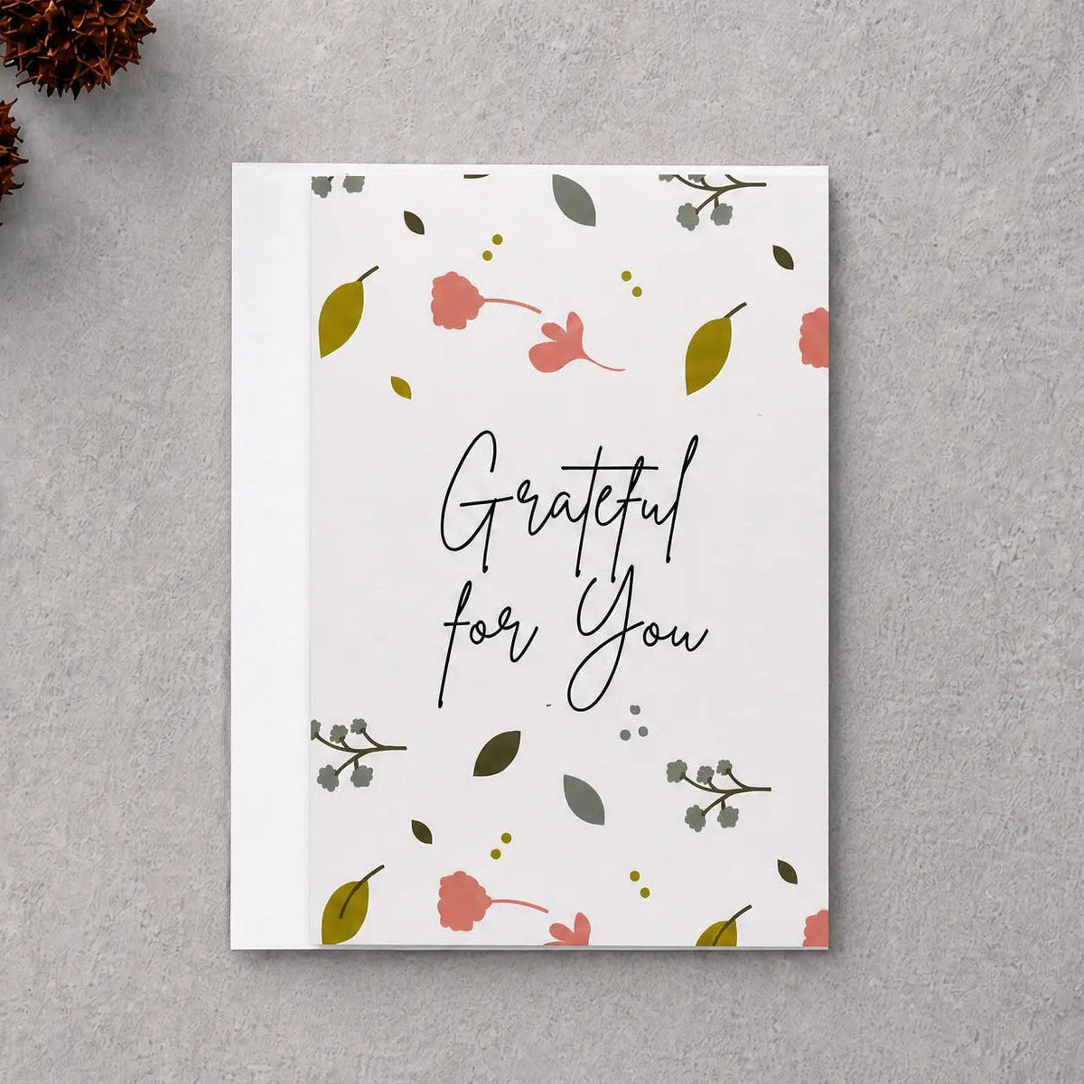 A square white greeting card with the phrase 'Grateful for You' in black script, decorated with small pink flowers, green leaves, and delicate botanical illustrations. The card is displayed in a flat-lay arrangement on a textured gray background, surrounded by natural decorative props including dried brown leaves, pinecones, and a piece of driftwood.