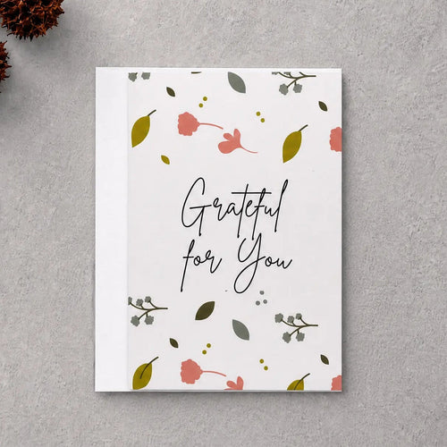 A square white greeting card with the phrase 'Grateful for You' in black script, decorated with small pink flowers, green leaves, and delicate botanical illustrations. The card is displayed in a flat-lay arrangement on a textured gray background, surrounded by natural decorative props including dried brown leaves, pinecones, and a piece of driftwood.